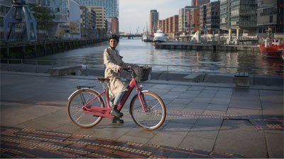 Zu sehen ist eine Frau auf einem rosa Cube Ella Hybrid E-Bike. Sie fährt an einem Fluss entlang. Im Hintergrund ist eine Stadt.
