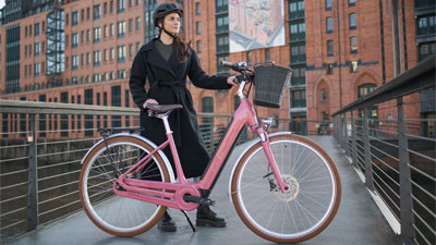 Zu sehen ist eine Frau hinter einem rosa Cube Ella Hybrid E-Bike. Im Hintergrund ist ein Gebäude.