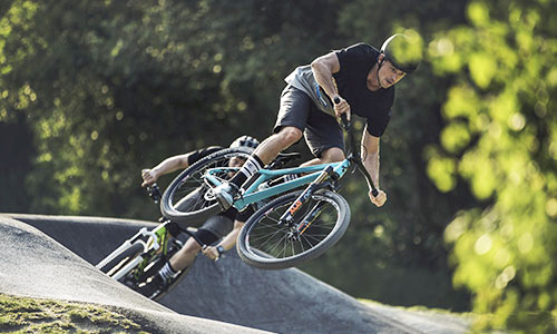 Radfahrer springt mit einem türkisblauen Dirtbike in einem Bikepark, im Hintergrund ist ein weiterer Radfahrer mit grünem Fahrrad zu sehen.