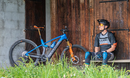 Zu sehen ist ein Radfahrer, der auf einer Bank vor einer Scheune sitzt. Neben ihm steht sein Fully E-Mountainbike.