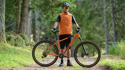Ein Mann steht hinter seinem orangen Cube Mountainbike, auf einem Waldweg.