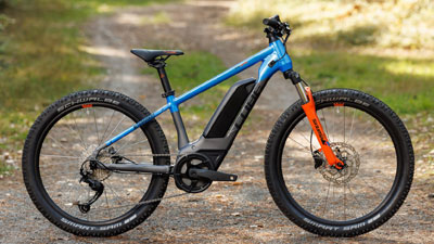 Zu sehen ist ein graues Cube Kinder E-Bike, mit blauen und orangen Details. Es steht auf einem Waldweg.