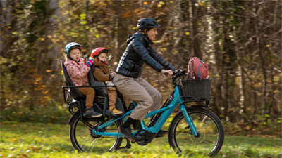 Zu sehen ist eine Mutter auf einem blauen Cube Longtail Lastenrad. Auf dem Gepäckträger sitzen zwei Kleinkinder.